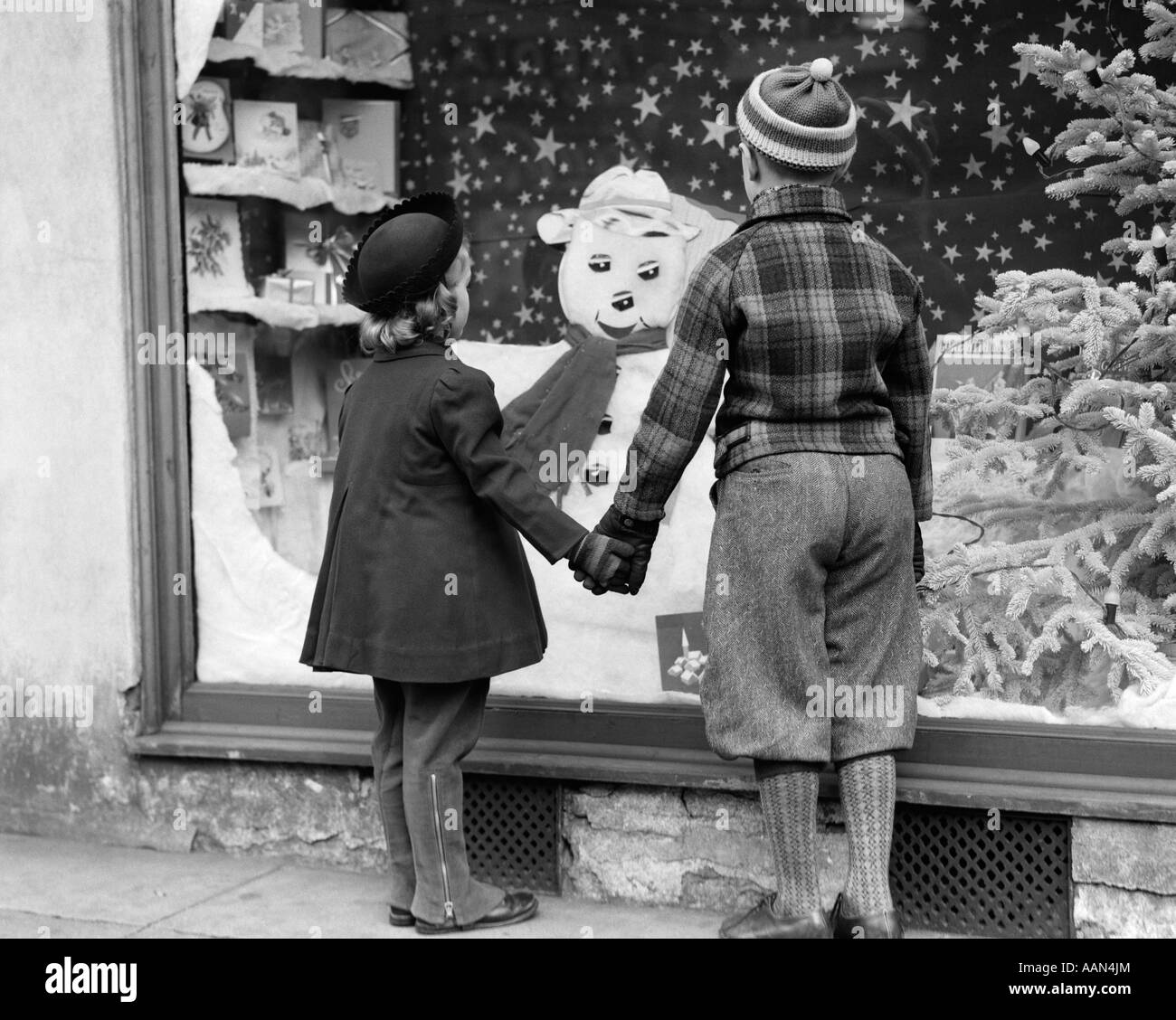 1930s BACK VIEW BOY AND GIRL HOLDING HANDS LOOKING AT DECORATED CHRISTMAS WINDOW Stock Photo