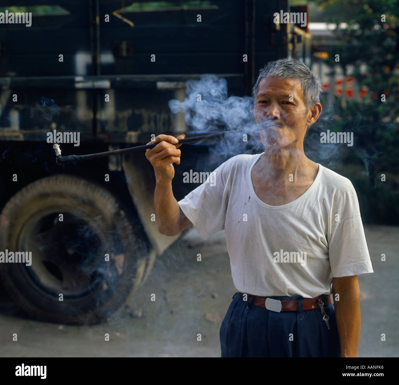 Elderly man smoking traditional pipe with long stem with old truck in background in Chongqing Sichuan China Stock Photo