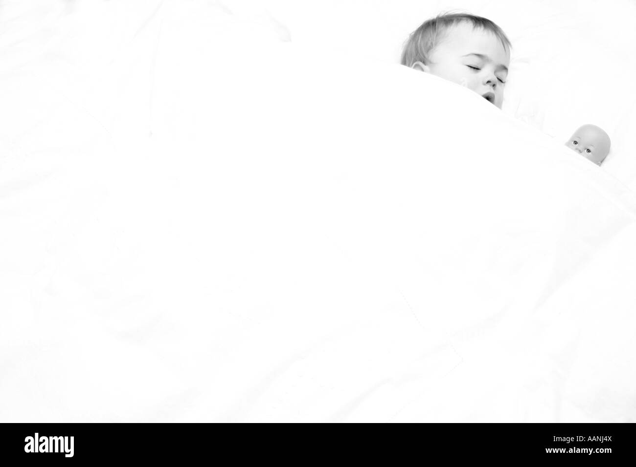 high key black and white shot of 18 month old girl asleep in a bed with her doll under white duvet Stock Photo