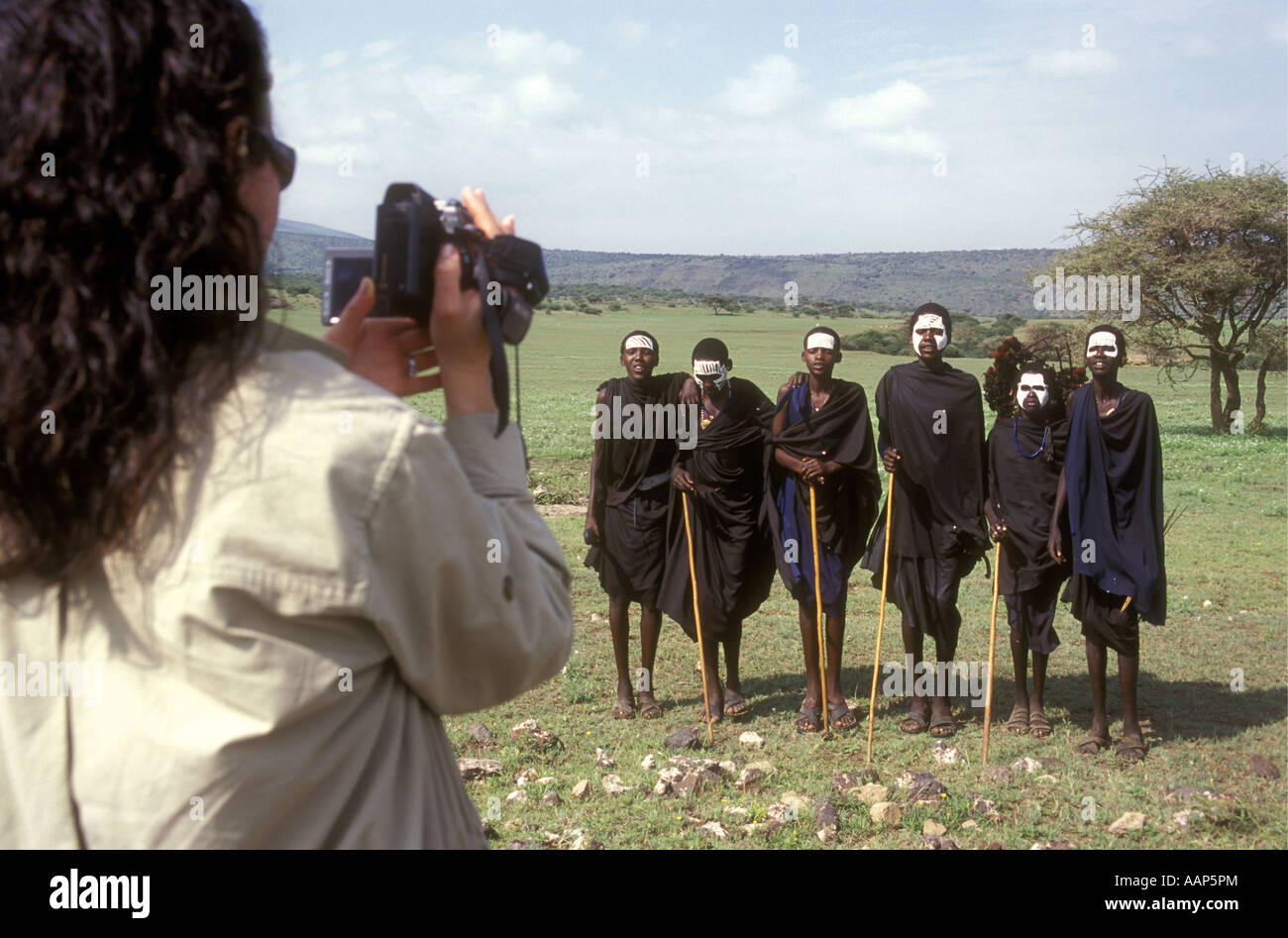 Six young Maasai men with black cloaks and white face paint being filmed by a white tourist Tanzania Stock Photo