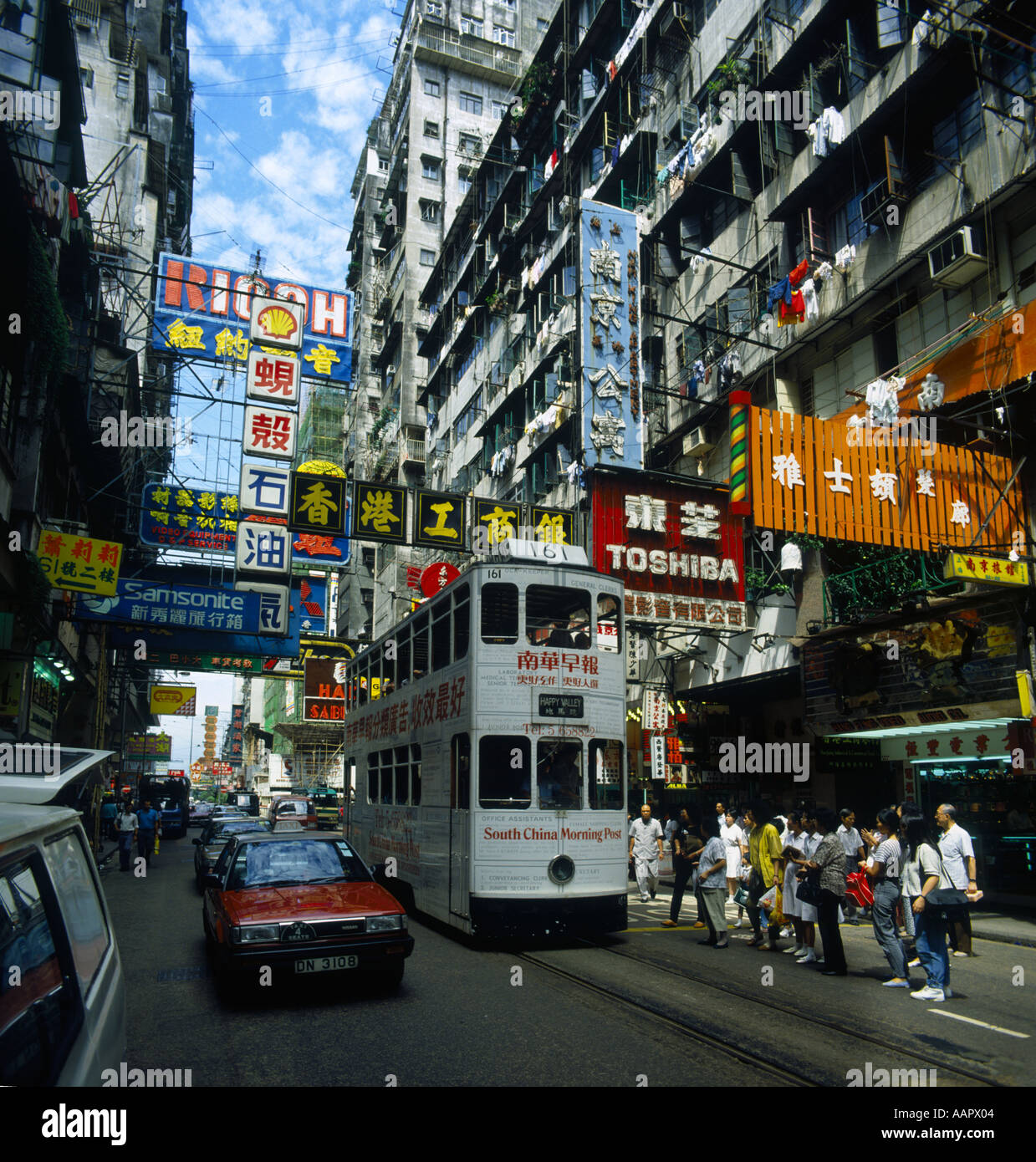 Busy narrow street scene in Wanchai District people waiting for tram red taxi on left many Chinese signs overhead in Hong Kong Stock Photo