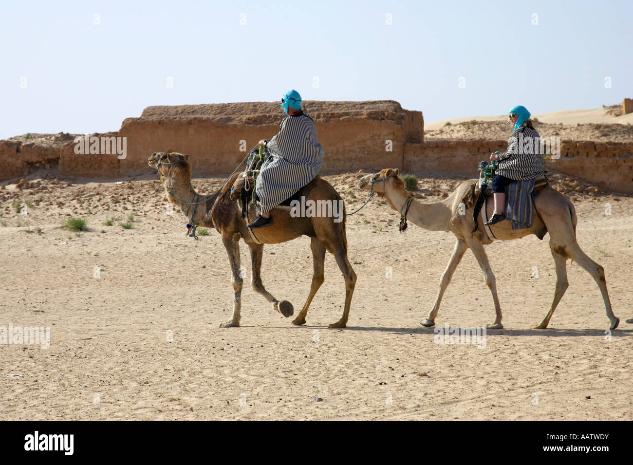 tourists in desert clothing ride camels through ghost town in the sahara desert at Douz Tunisia Stock Photo