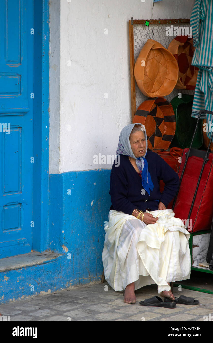 vertical old tired local woman in headscarf rests weary feet beside market stall and traditional blue door nabeul tunisia Stock Photo