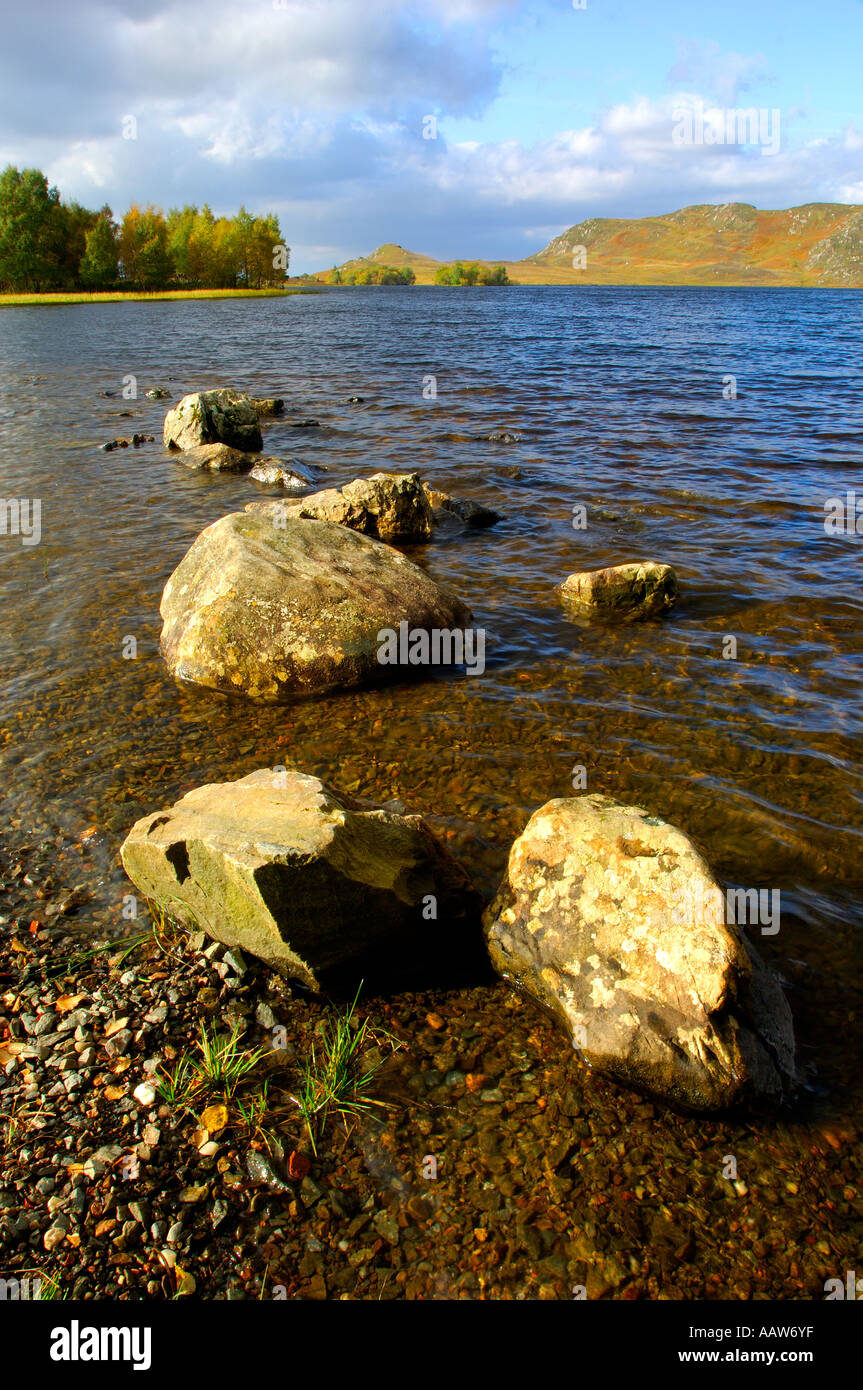 Tranquil image of Loch Tarff in the Scottish highlands with rocks leading into the distance towards a mountainous backdrop Stock Photo