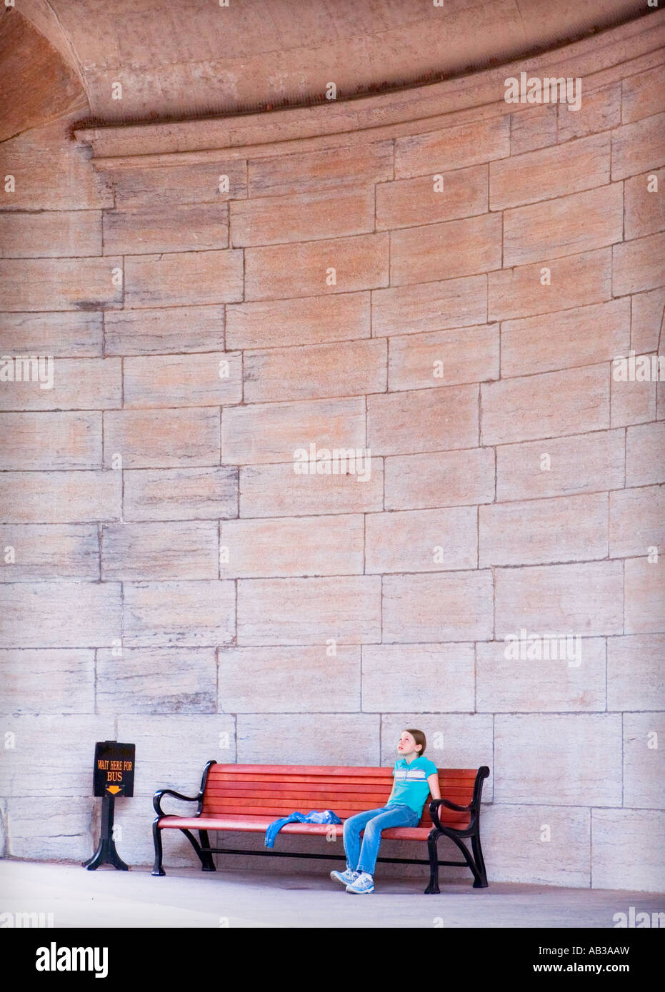 female child sits on a wooden bench by a tall stone wall waiting for a bus and looking up Stock Photo