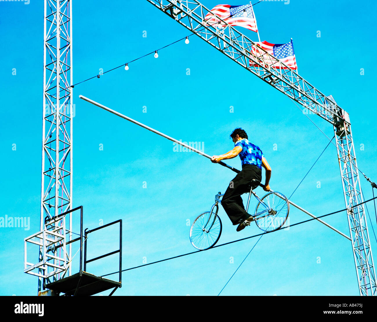 high wire walker riding bicycle set against blue sky with American flags flying overhead Stock Photo