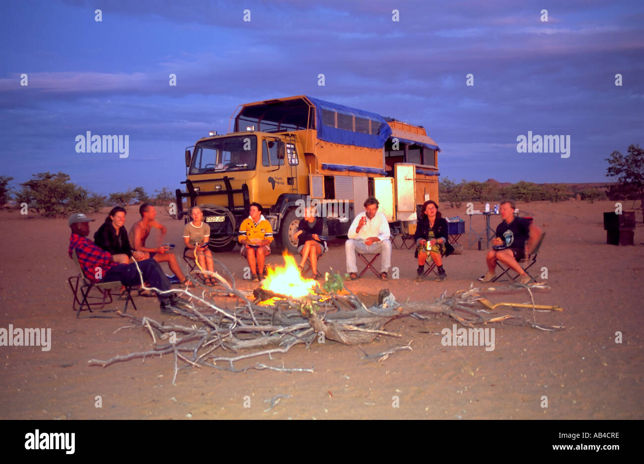 Tourists sitting round a fire at a bush campsite on an overland trip in the Namibian desert. Stock Photo