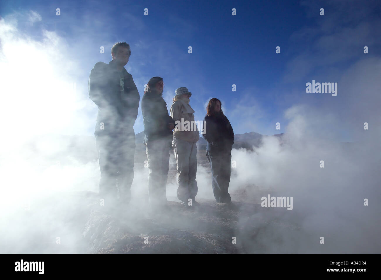 Four tourists semi silhouetted among the steam from the geysers at El Tatio in the early morning sun. Stock Photo