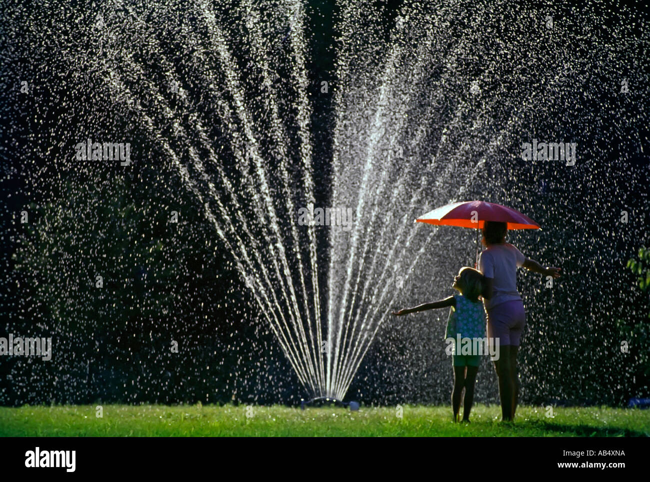 Mother with a red umbrella and 4 year old daughter stand under a lawn sprinkler and catch water drops Stock Photo