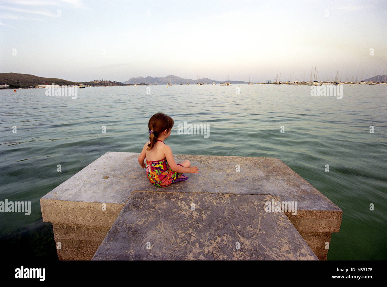 A small girl sits on the end of a jetty in the early morning in Puerto Pollensa in Majorca Stock Photo