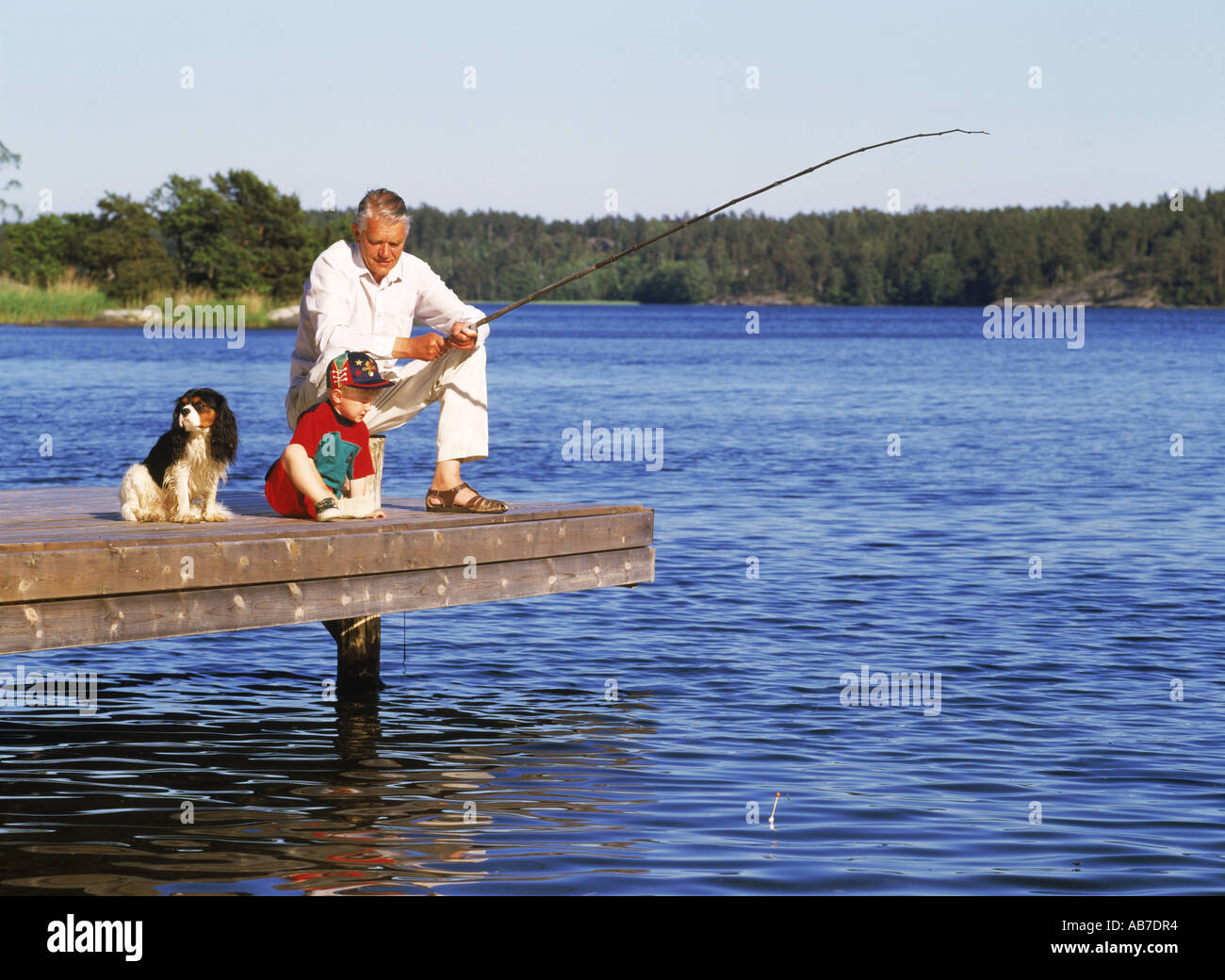 Granddad fishing with grandchild and family dog on lakeside pier during Swedish summer Stock Photo
