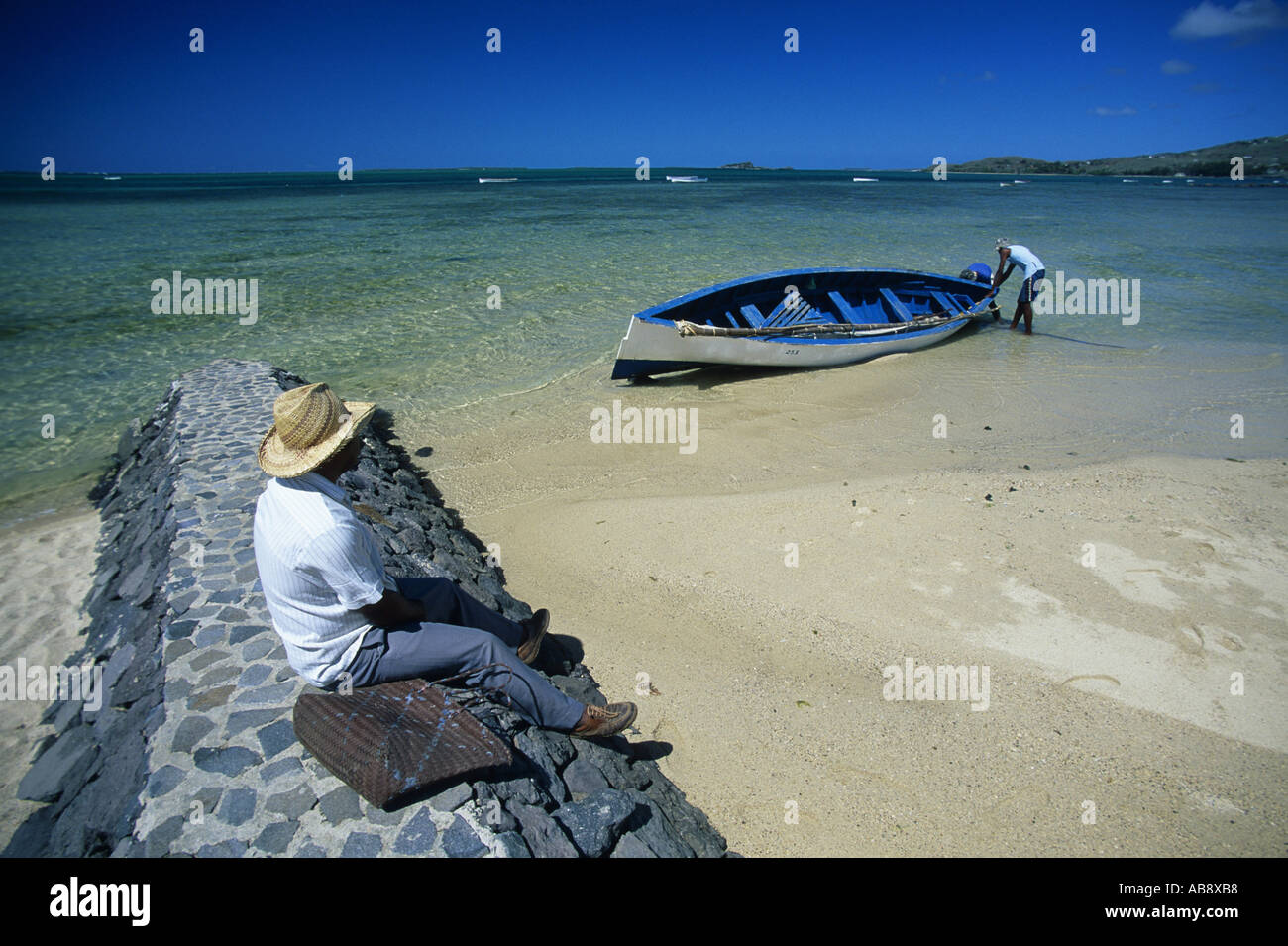 three men at a tropical beach, one sitting on a breakwater with a bag, the others launching a boat, Mauritius, Rodrigues. Stock Photo
