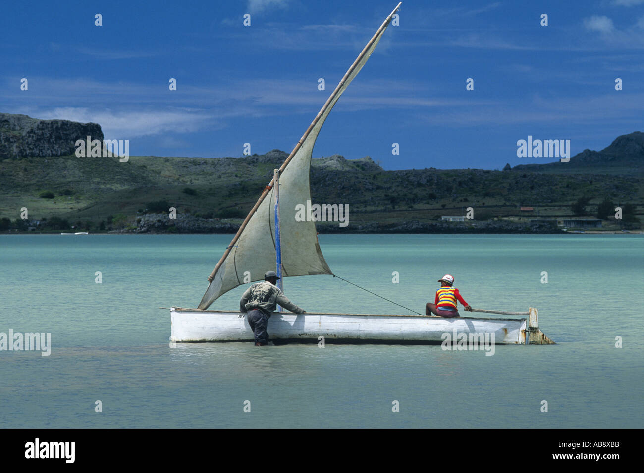 two men in tropical bay with sailing boat on sand bank, one standing in water trying to free boat, the other navigating, Maurit Stock Photo