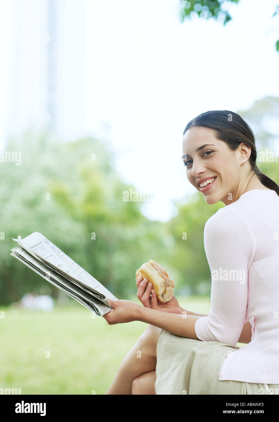 Young woman reading newspaper and eating sandwich in city park Stock Photo