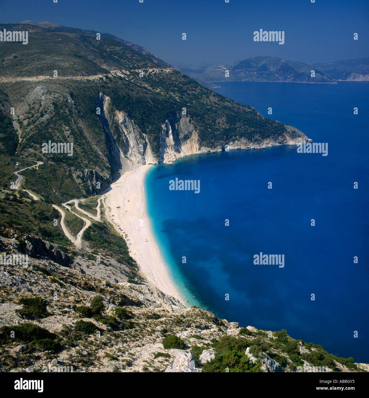 Looking down on bay with clear blue water white sand beach and zig-zag road down hillside Mirtos Cephalonia Island Greek Islands Stock Photo
