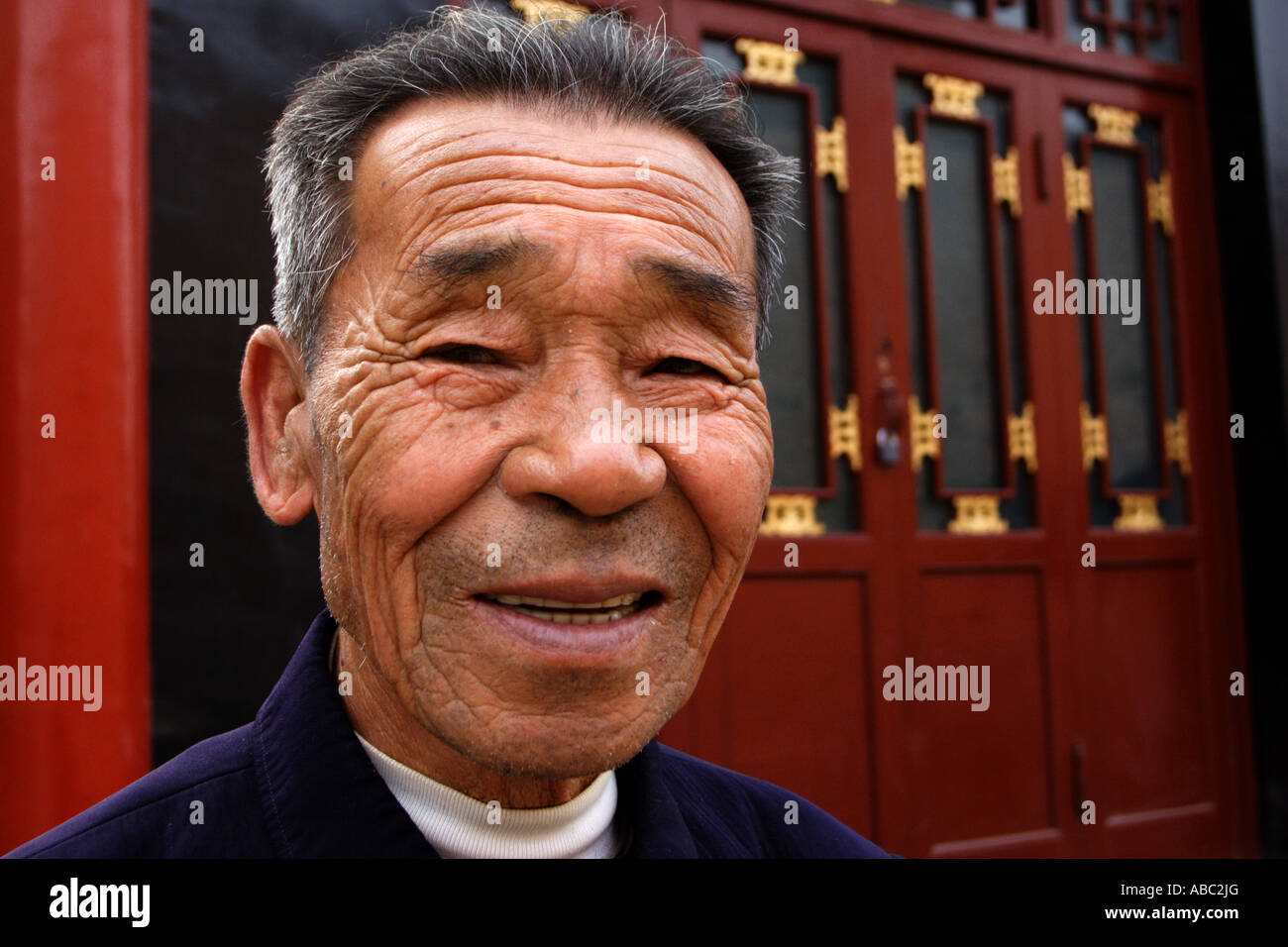 An elderly man in Pingyao, China, grins. Behind him stands a traditional red laquer facaded Chinese shop. Stock Photo
