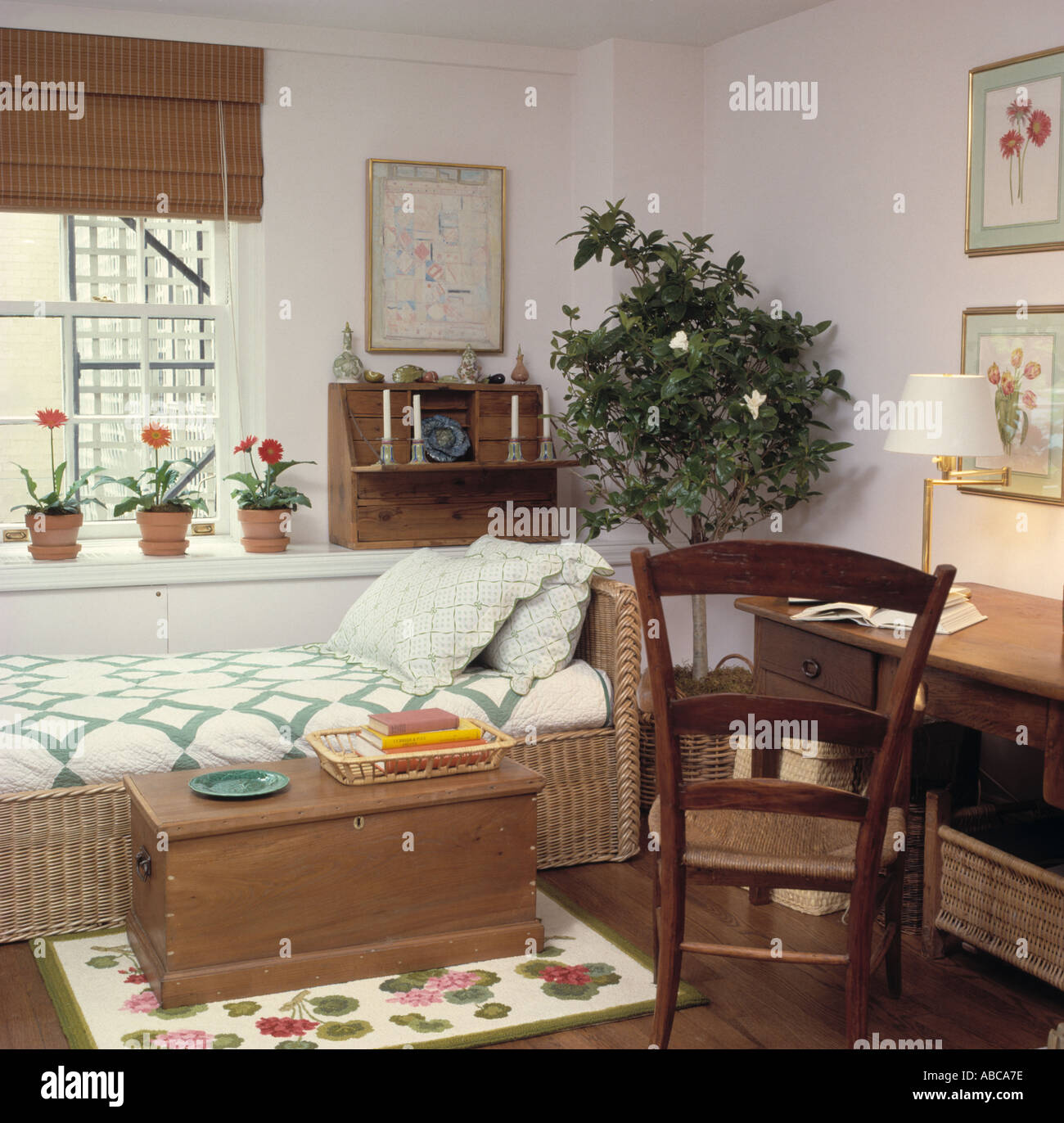Ladder-back chair and wooden chest in bedroom with patchwork quilt on wicker bed in front of window with split-cane blind Stock Photo