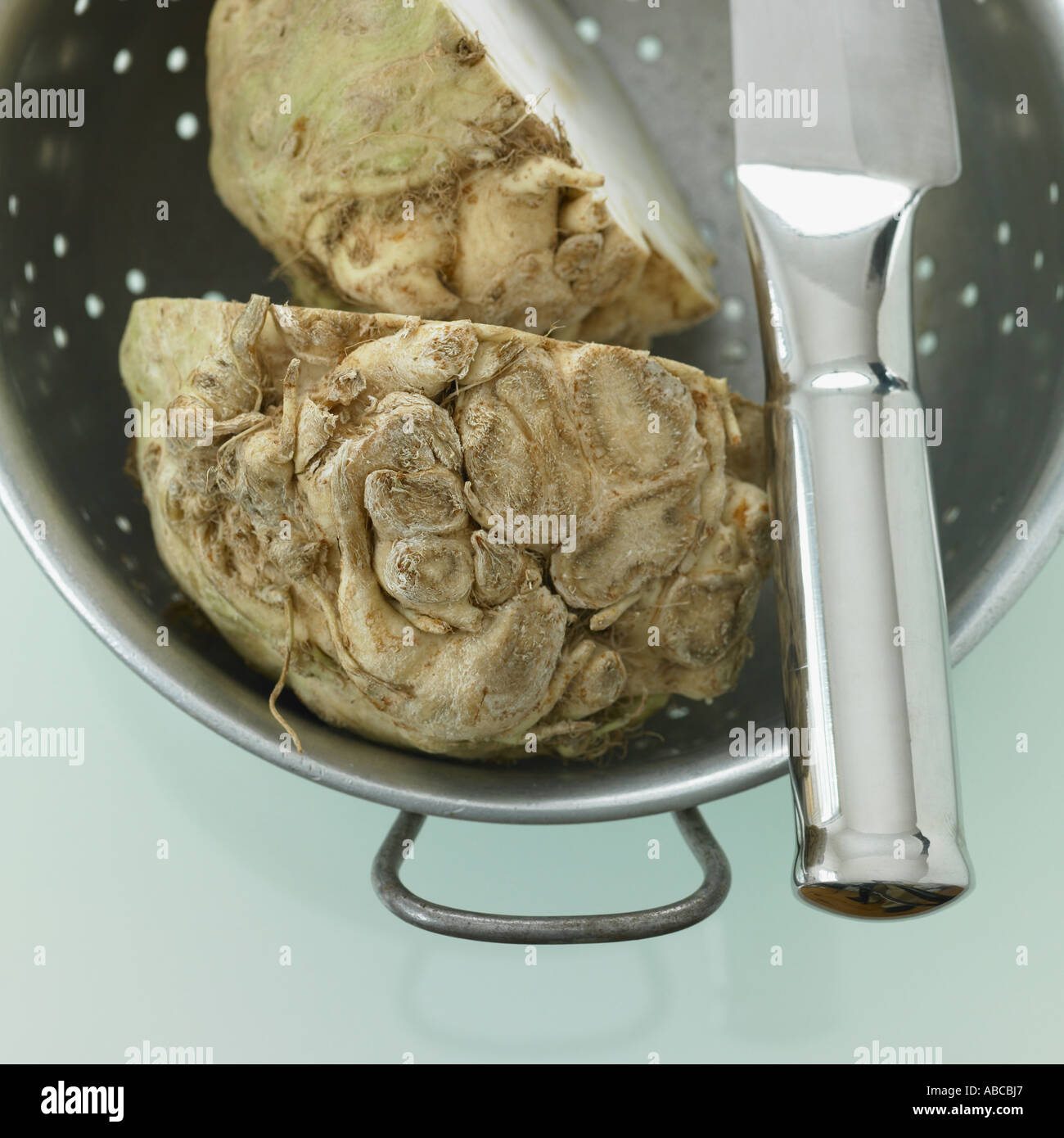 Halved celeriac in colander with stainless steel knife on pale green glass background Stock Photo