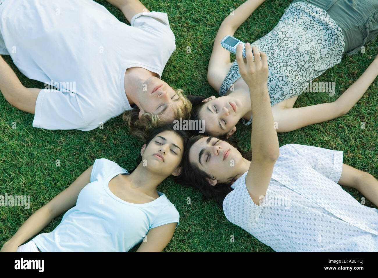 Group of young friends lying on grass with heads together, eyes closed, one taking photo with cell phone Stock Photo