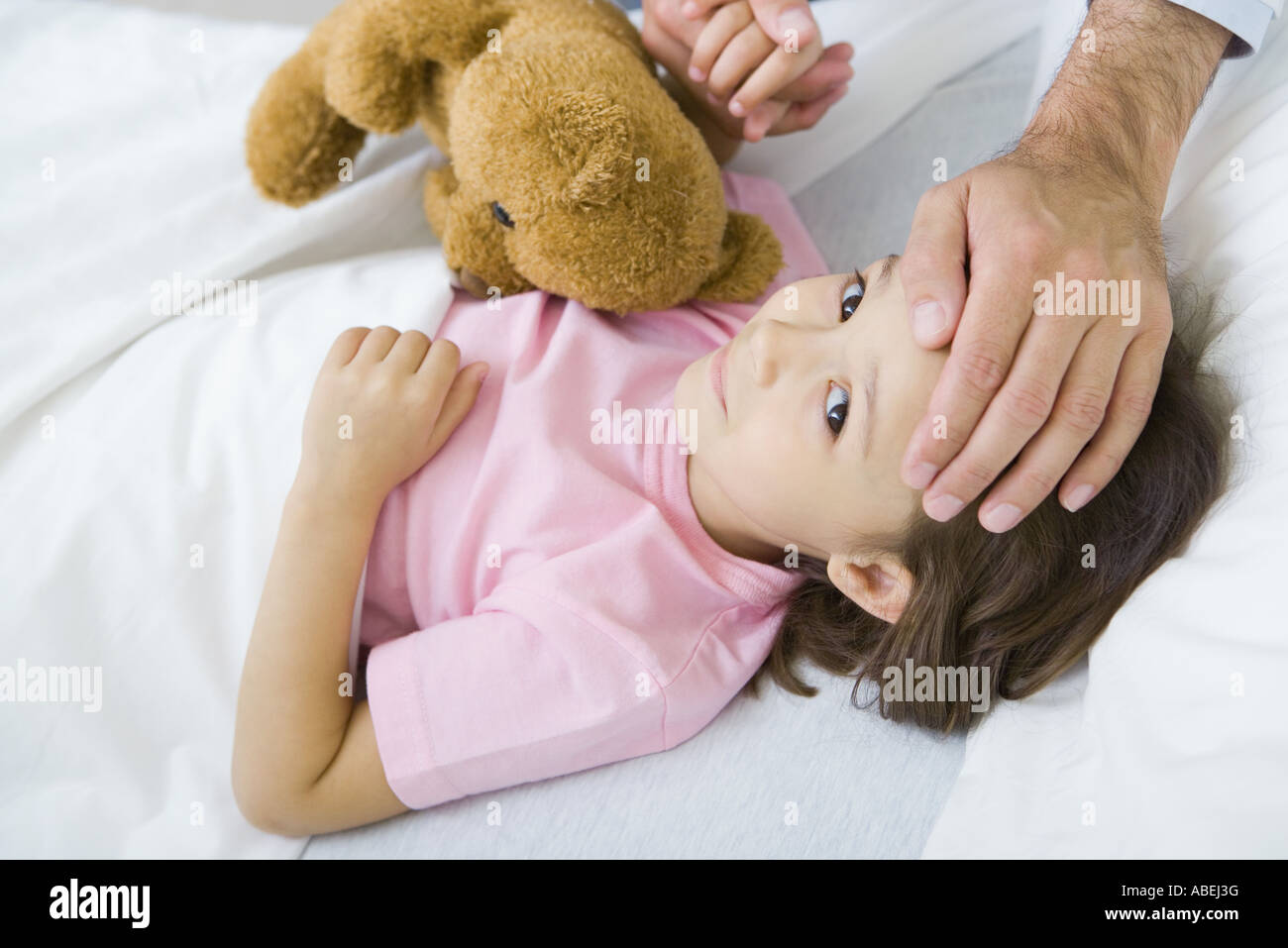 Girl lying in bed with teddy bear, man holding her hand on putting other hand on her forehead Stock Photo