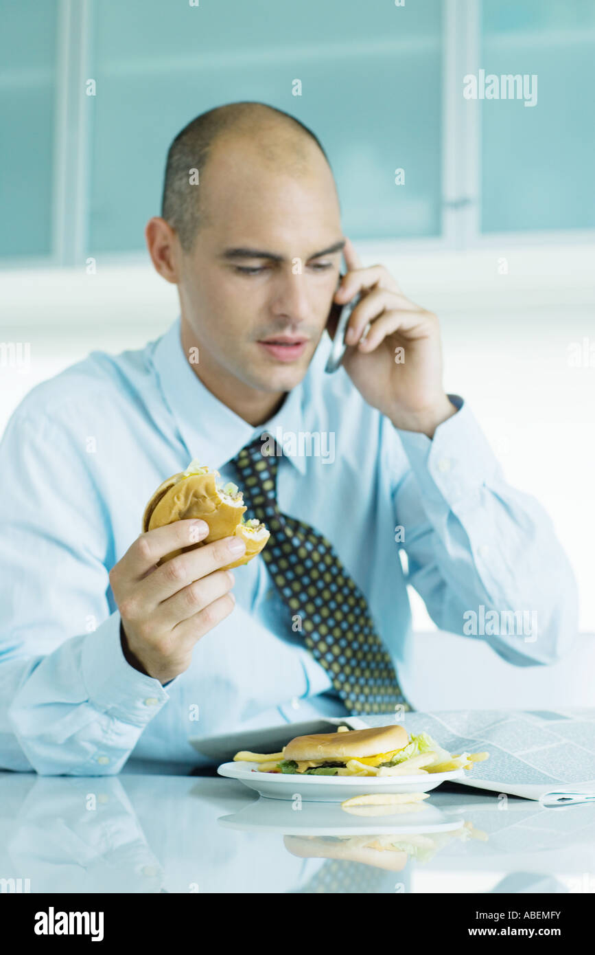 Man sitting at table eating hamburgers, reading newspaper and using cell phone Stock Photo