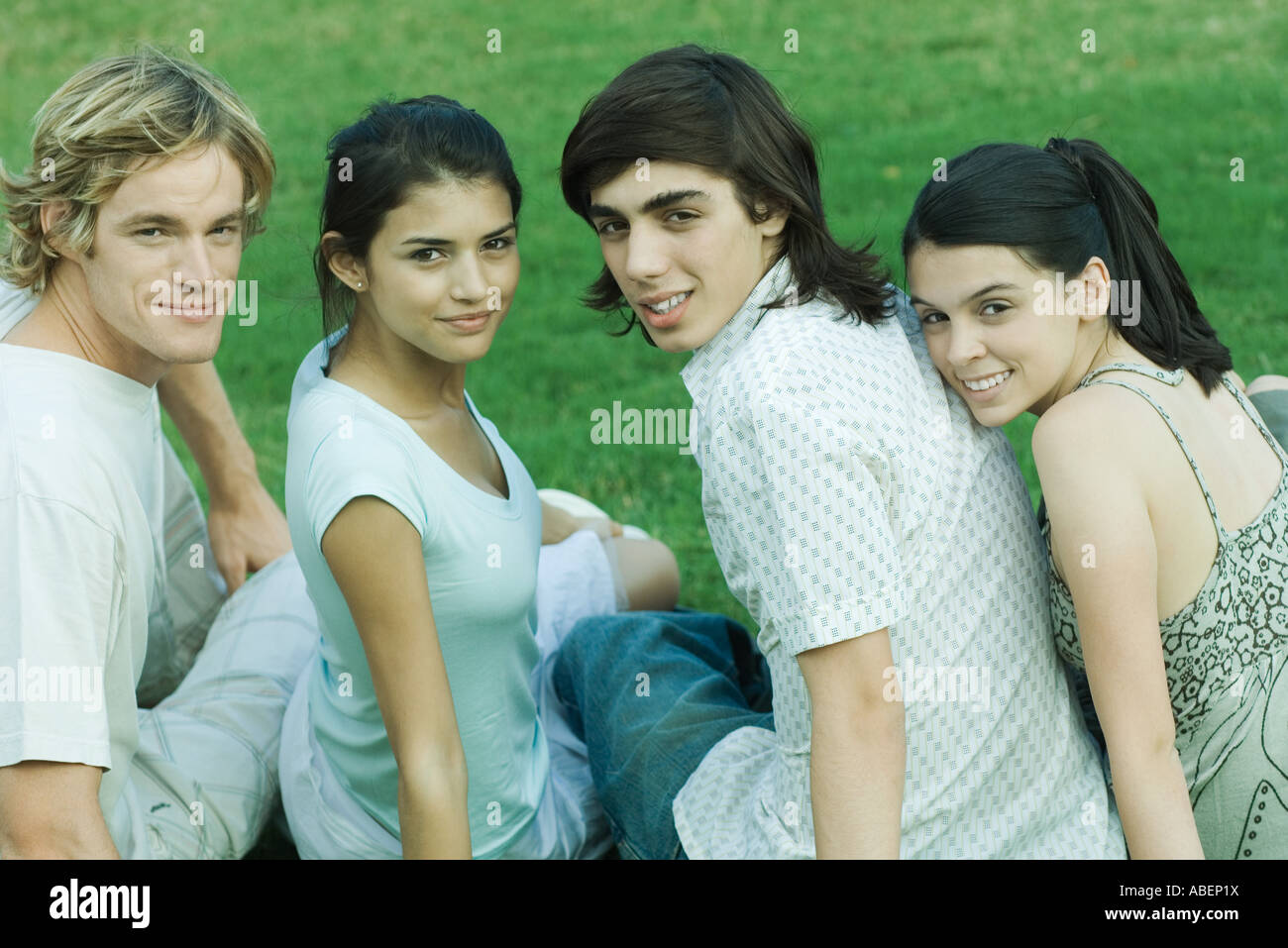 Four young friends sitting on grass, side by side, looking over shoulders at camera Stock Photo