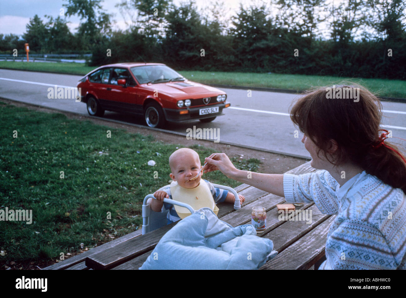 Mother spoon feeding her baby son in a layby with a red Alfa Romeo GTV6 sports coupe in the background Stock Photo