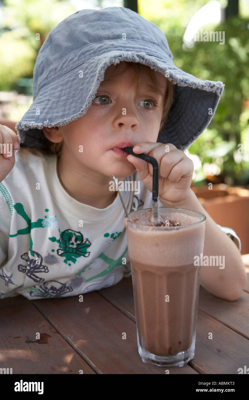 young child in summer wearing floppy sun hat at a cafe having a large glass of iced chocolate drinking through a straw Stock Photo