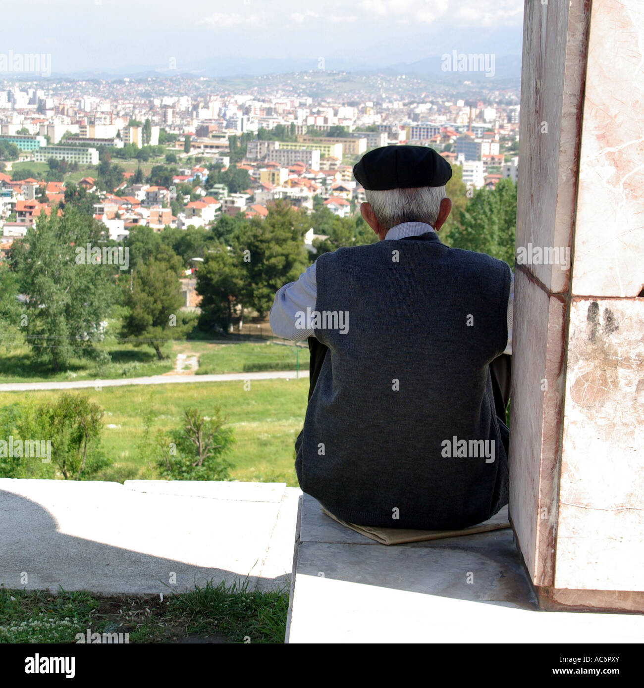 City of Tirana in Republic of Albania sprawls out below an old man sitting on wall overlooking the urban landscape from hillside in formal parkland Stock Photo