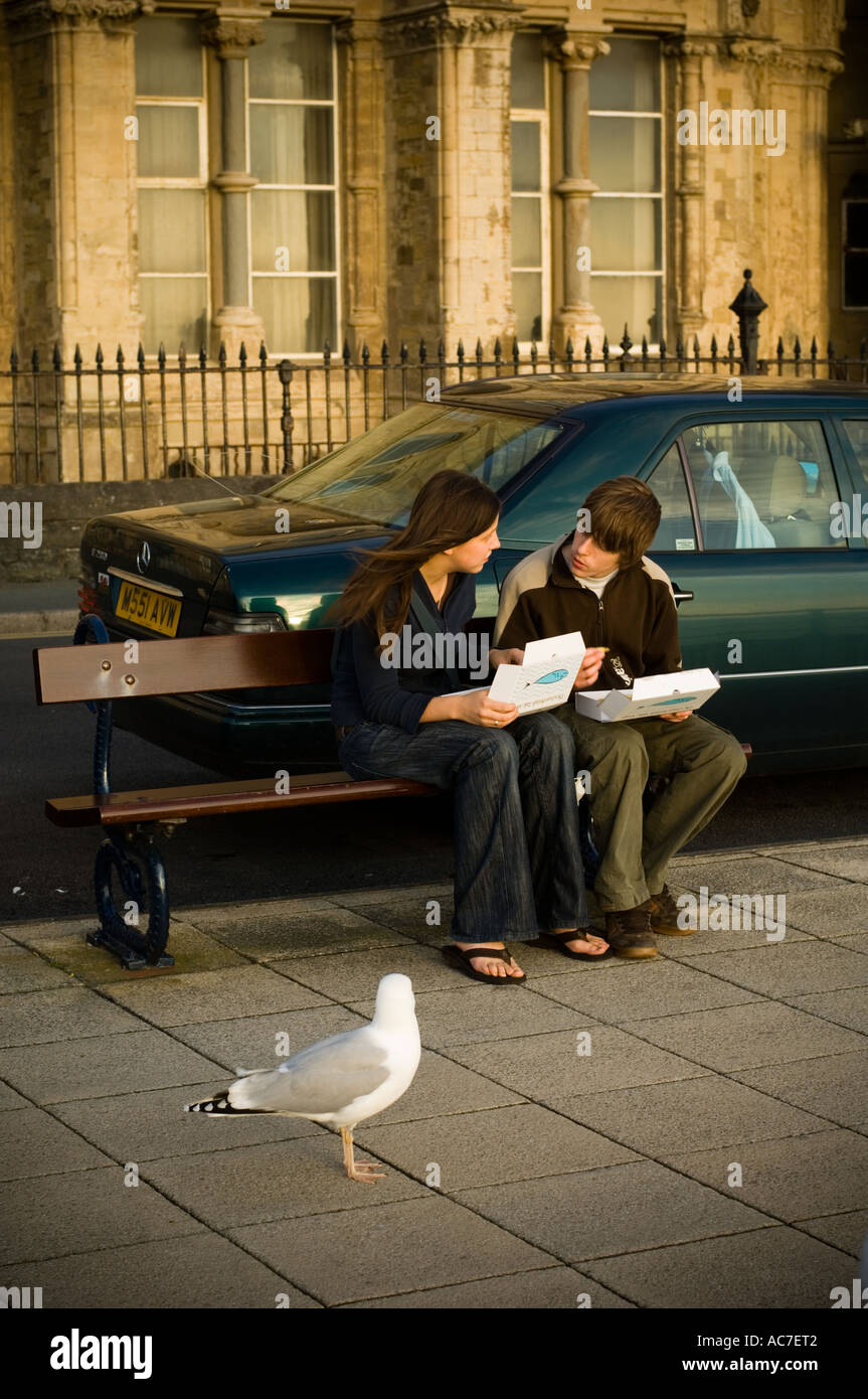Young couple eating fish and chips sitting on Aberystwyth promenade while a seagull looks on Stock Photo