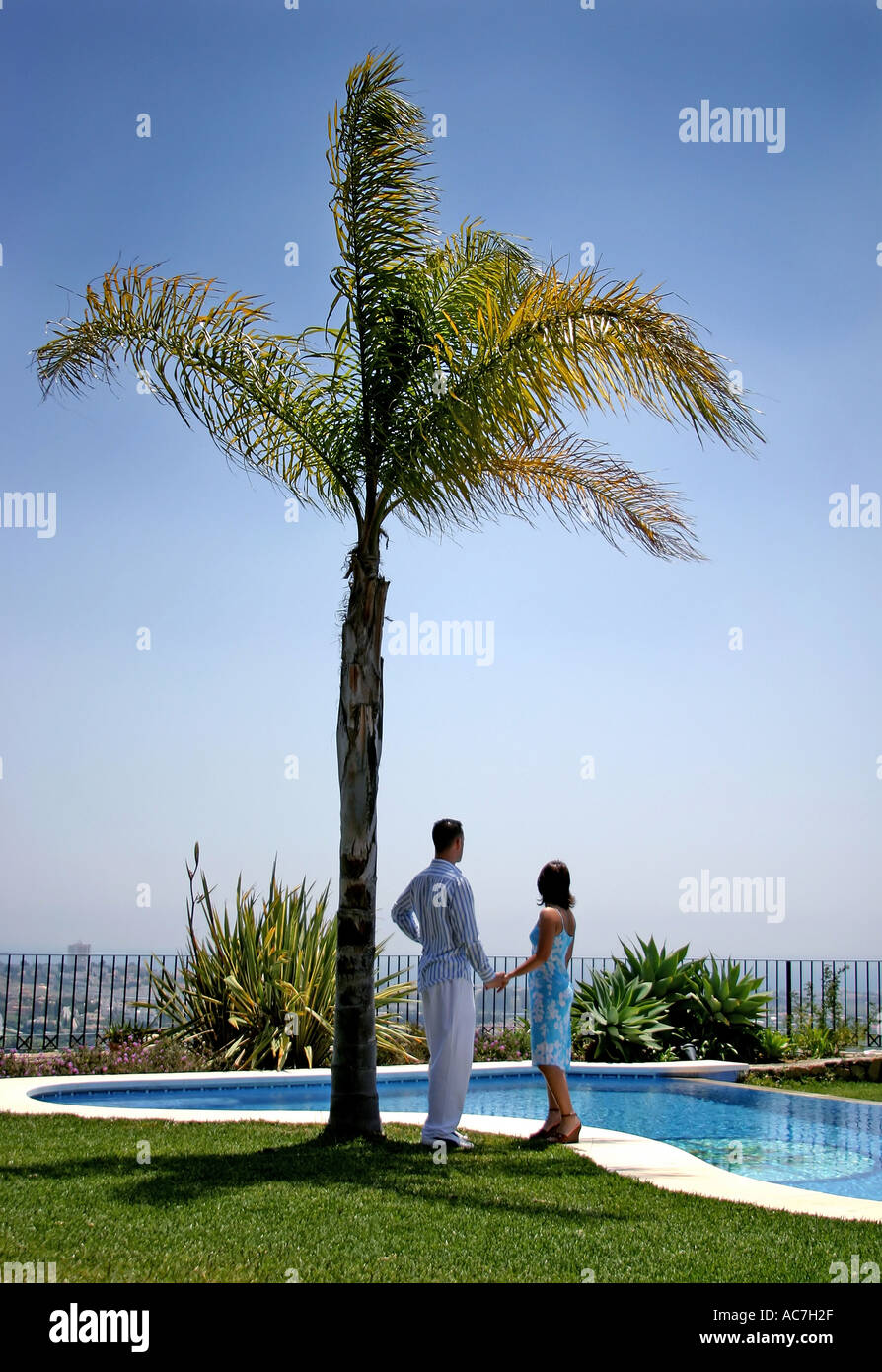 Couple holding hands under a palm tree in the sun in Spain next to the swimming pool  Stock Photo