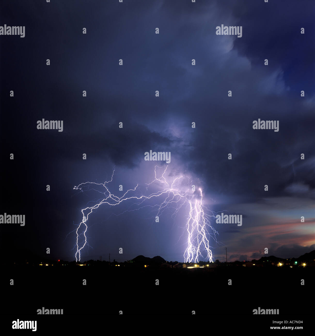 Lightning strikes over a city dotted with glowing lights from a dark blue stormy sky in Southern Arizona during a monsoon storm. Stock Photo