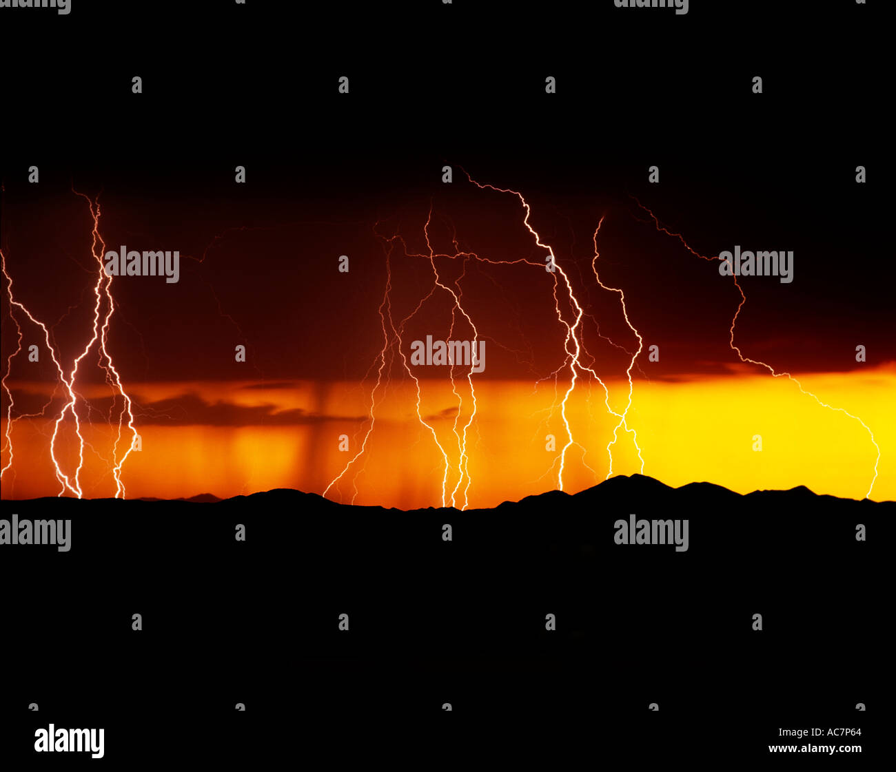 Lightning strikes during a vibrant orange and yellow sunset over mountains west of Tucson, AZ, USA. Rain falls behind the bolts. Stock Photo