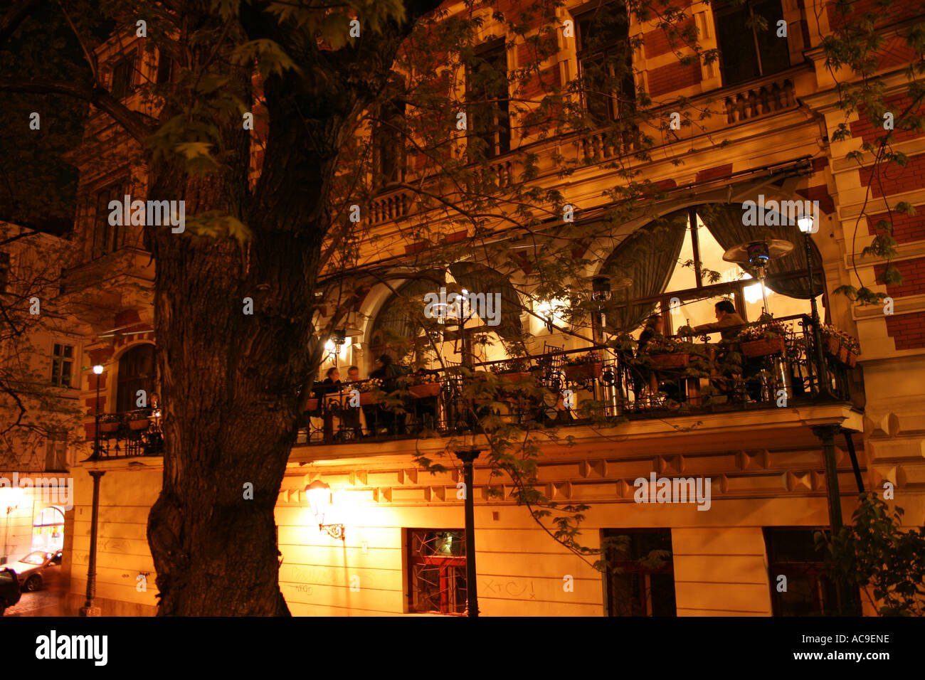 Outdoor restaurant on a charming balcony of a historic building in Prague, illuminated at night, with people dining and enjoying the ambiance. Stock Photo
