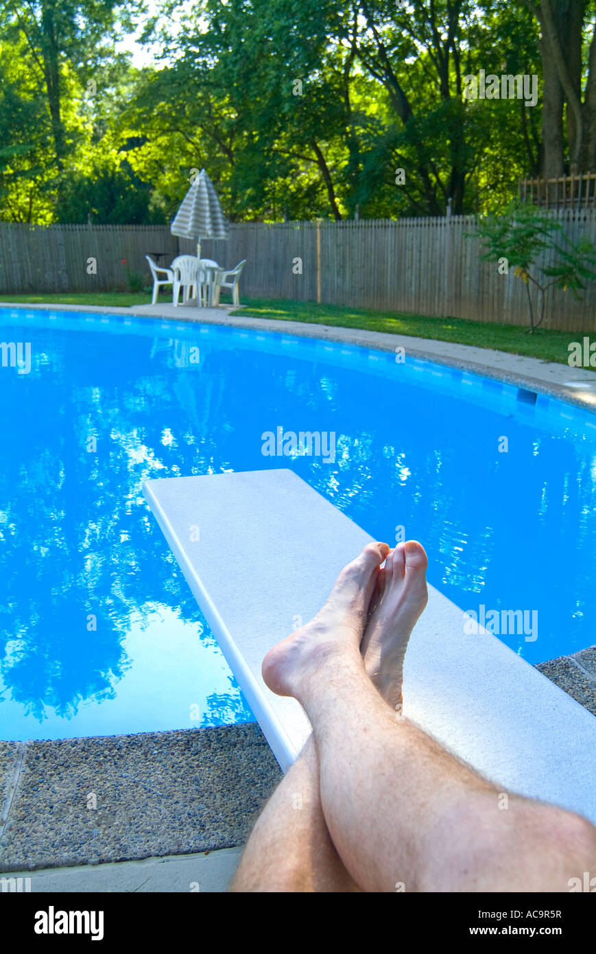 Swimming Pool With Mans Tan Legs On Diving Board Relaxing On A Summer Vacation Day Stock Photo