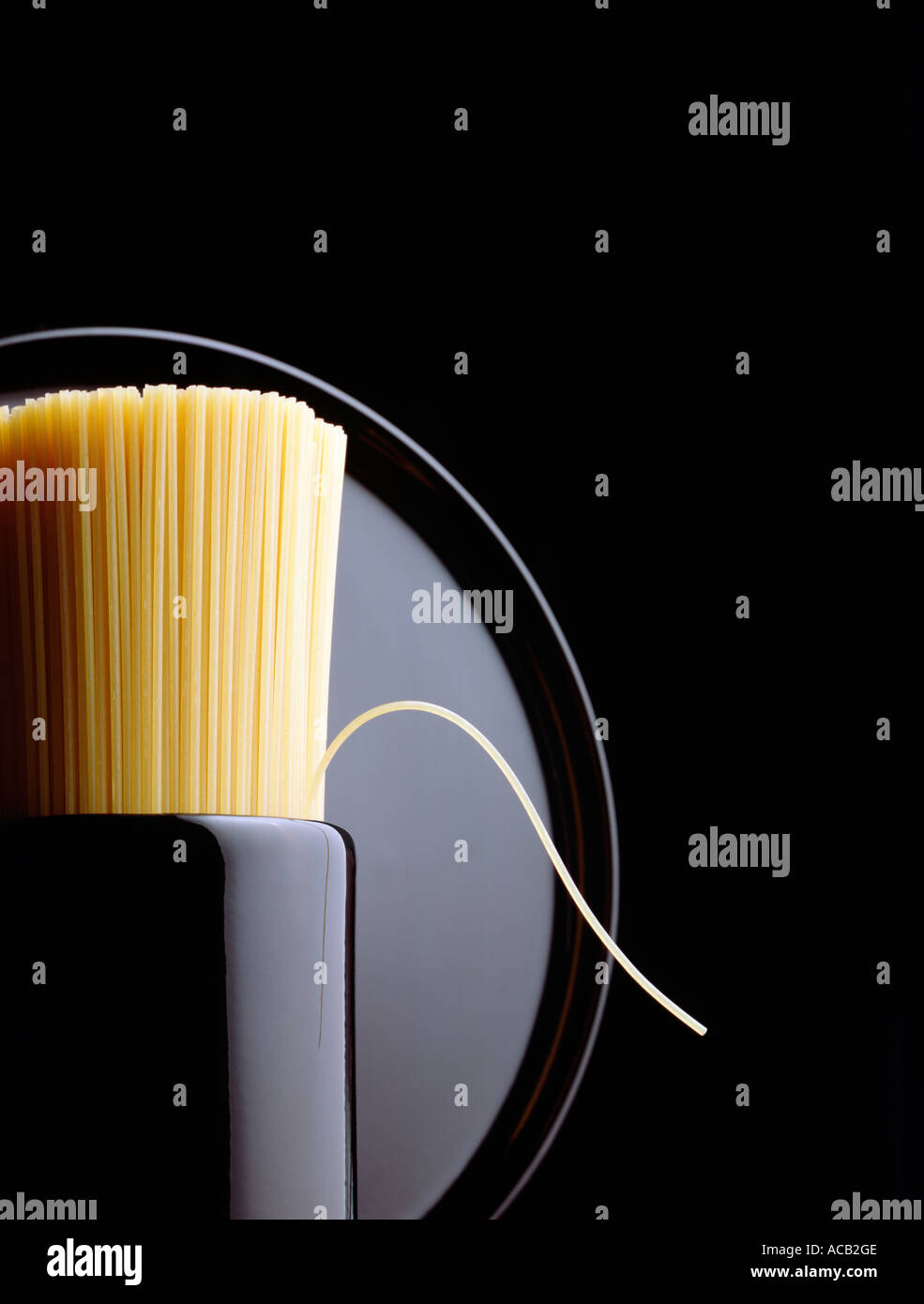 Spaghetti in black ceramic container one strand bent over plate behind Stock Photo