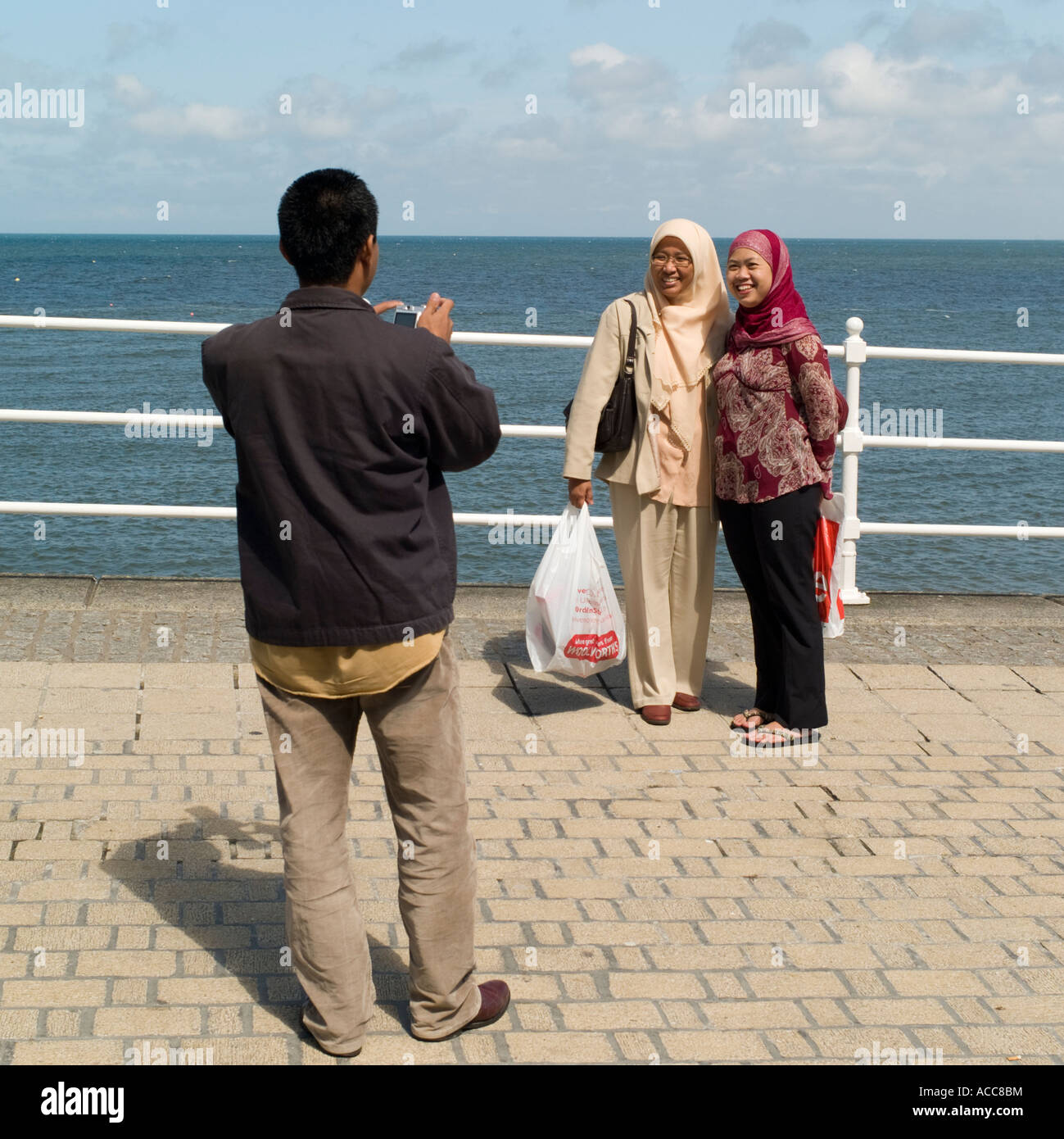 two happy smiling Muslim women being photographed by a male friend standing on aberystwyth promenade, summer afternoon Wales UK Stock Photo