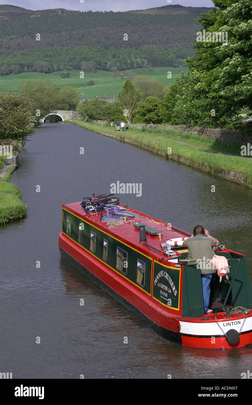 Red & green narrowboat (hire boat) sailing on scenic rural Leeds Liverpool Canal on sunny day, 1 man standing by tiller - North Yorkshire, England, UK Stock Photo