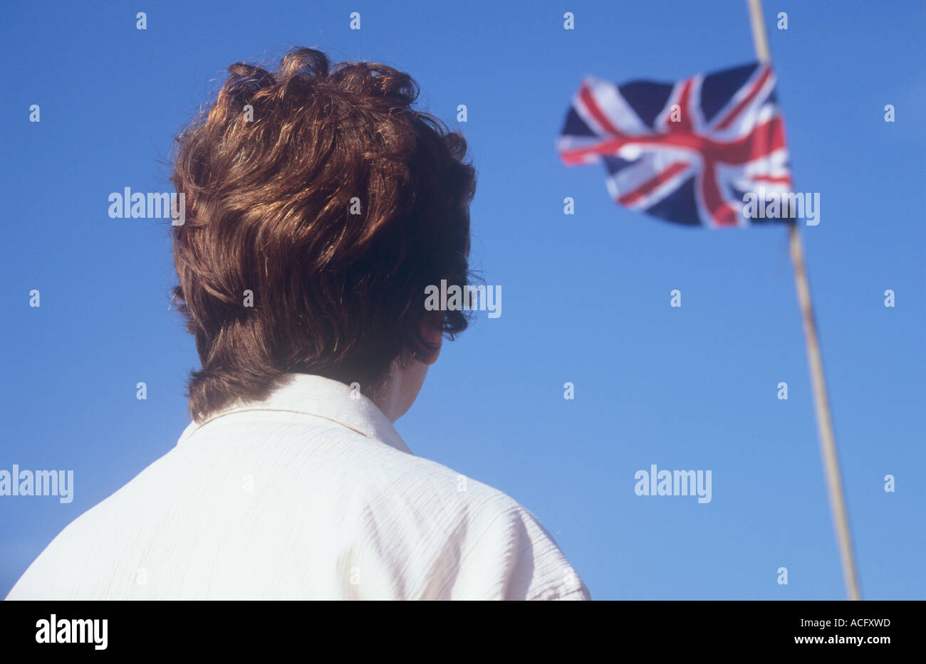 Backview of head of female figure in cream shirt gazing up at a distant Union Jack flag fluttering in a strong breeze Stock Photo