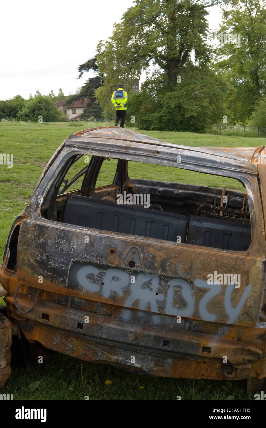 Burned vehicle covered in graffiti abandoned in a field and police officer patrolling the area Stock Photo
