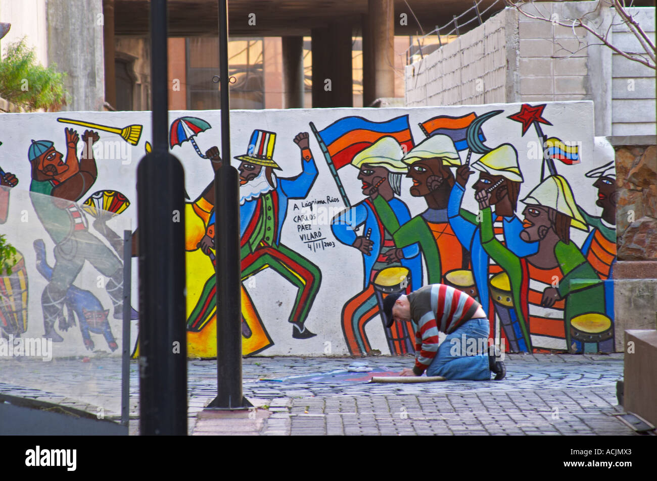 A man making a street painting on the pavement in front of a wall fresco painting with colourful men depicting an historic event titled A Lagrima Rios by Carlos Paez Vilaro Montevideo, Uruguay, South America Stock Photo