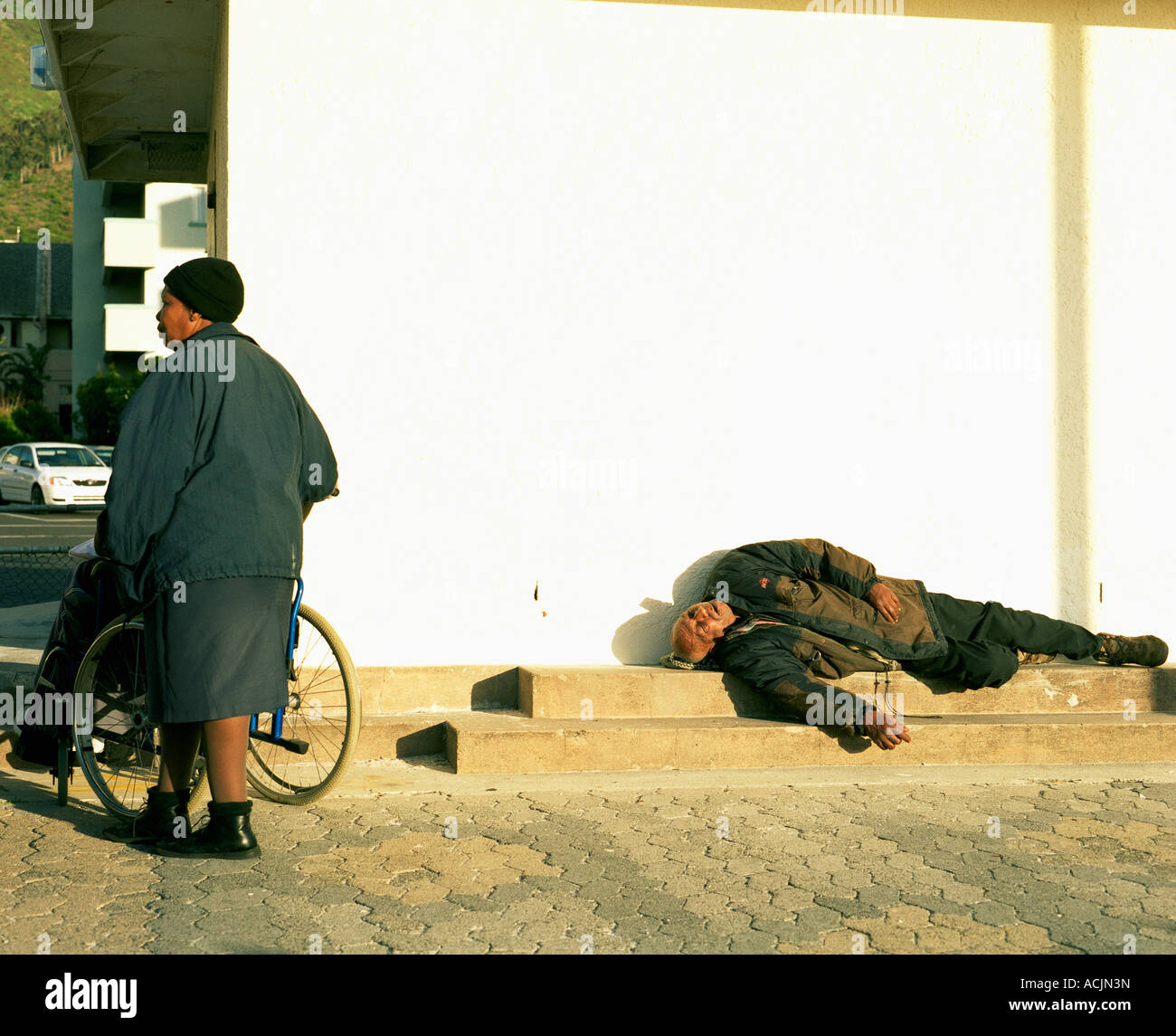 Woman pushing wheelchair while ignoring vagrant lying on ground in Cape Town Stock Photo