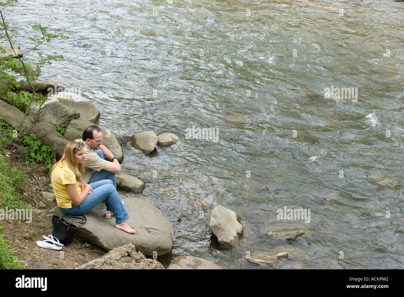 Young man and woman couple sitting peacfully on rock along the bank of the Little Pigeon River in downtown Gatlinburg, Tennessee, USA Stock Photo