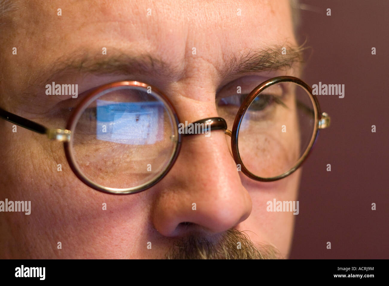 A computer screen reflects in a mustachioed man's circular glasses