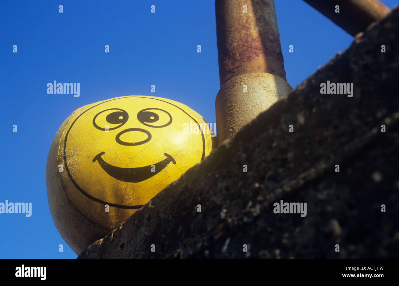 Soft padded yellow vinyl ball with cheeky smiley face and sand grains by rusty railings on concrete wall under clear blue sky Stock Photo