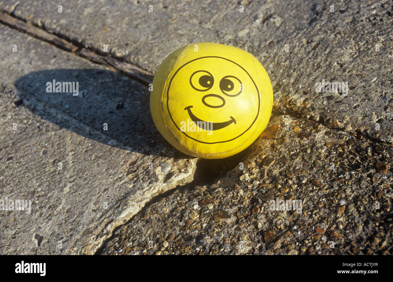 Soft padded yellow vinyl ball with cheeky smiley face and sand grains on concrete wall or path or ramp Stock Photo