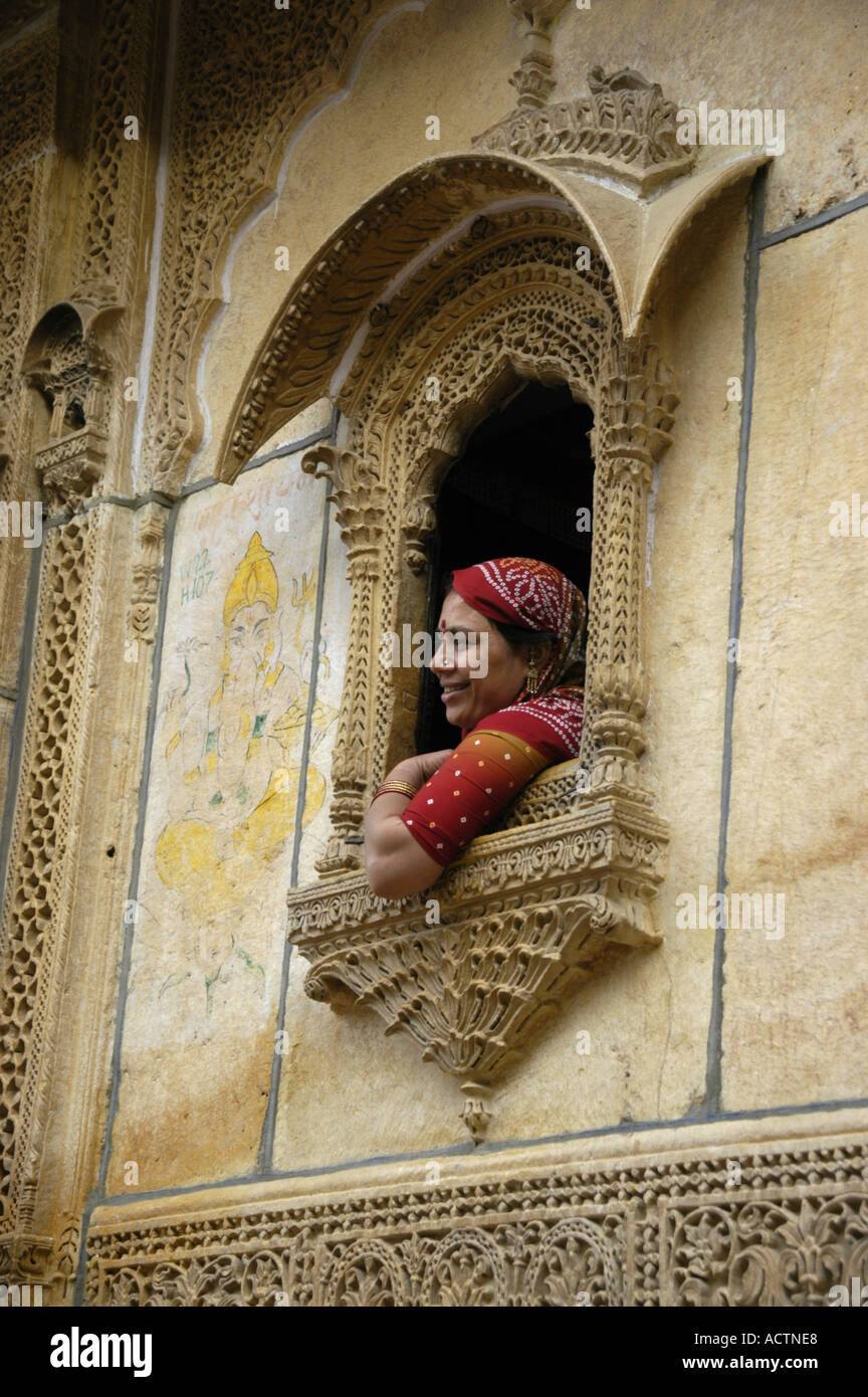 Indian woman dressed in red sari sits in a window decorated in fine arts of yellow sandstone Jaisalmer Rajasthan India Stock Photo