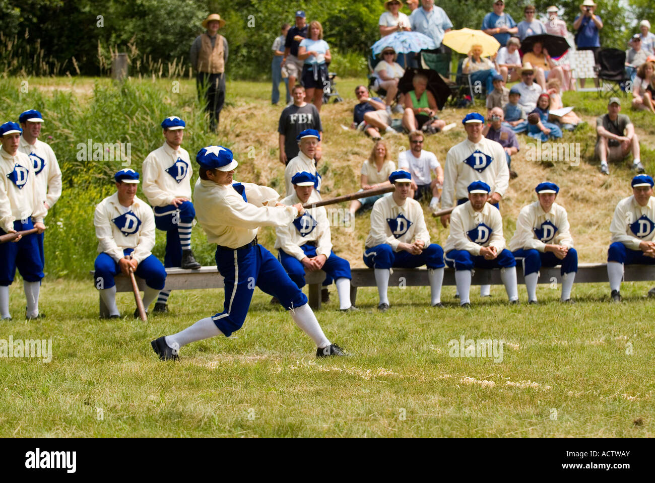 BASEBALL BATSMAN IS IN FULL SWING OF HIS BAT WHILE HIS TEAM MATES AND ONLOOKERS ARE WATCHING CLOSELY Stock Photo