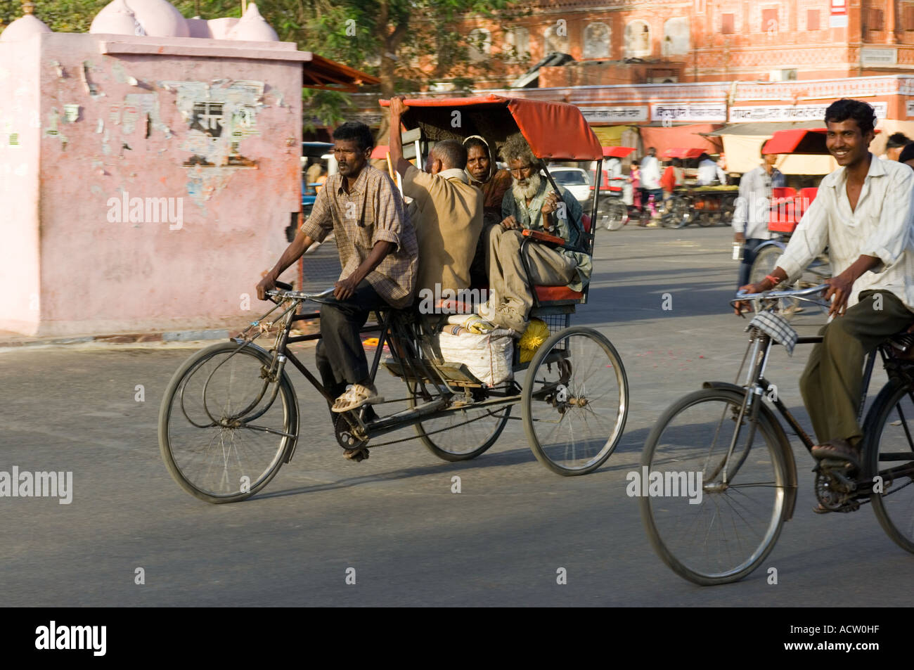 Several people squashed onto a cycle rickshaw and a man cycling in a typical street scene in Jaipur. Stock Photo