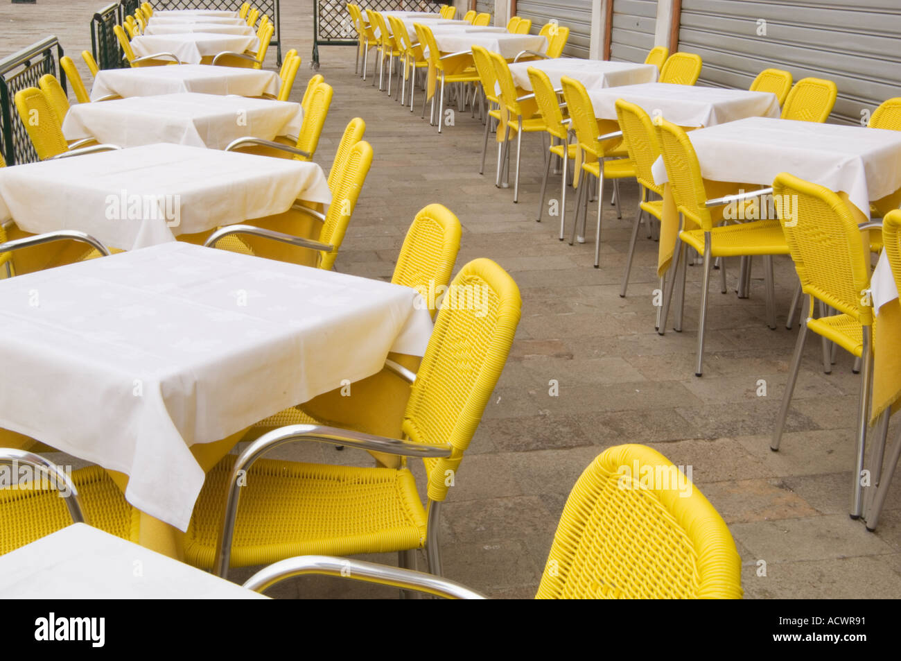 two rows of tables covered with white table cloths and surrounded by yellow wicker chairs in an outdoor cafe area in Venice Stock Photo